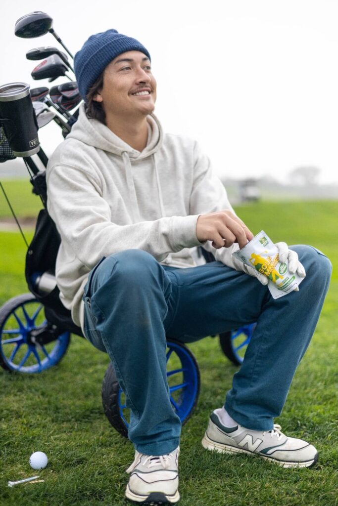 A person sits on a golf cart on a green field, holding a snack packet while wearing a grey hoodie and blue pants. Golf clubs are visible behind.